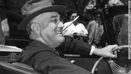 Dengan sebatang rokok di dudukan yang dikepalkan, giginya yang tersenyum, Franklin Delano Roosevelt duduk dengan gembira di atas roda mobil konvertibelnya, Warm Springs, Georgia, 1939. (Foto oleh Underwood Archives / Getty Images)